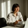 A woman sitting at her desk with a laptop in front of her, holding a credit card and smiling while looking out of the window.