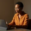 A woman with glasses working focused on her iPad connected to a keyboard, while holding an Apple Pencil.