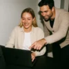 A woman working on her laptop and smiling while a man stands beside her, pointing at her screen in a modern office.