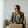 A professional woman smiling while seated on a sofa with a laptop in front of her.
