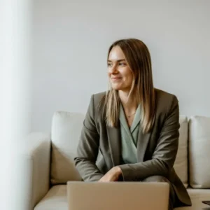A professional woman smiling while seated on a sofa with a laptop in front of her.