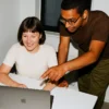 A smiling woman sitting at a desk while a man with glasses stands beside her, pointing at her laptop screen in a modern office.
