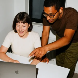 A smiling woman sitting at a desk while a man with glasses stands beside her, pointing at her laptop screen in a modern office.